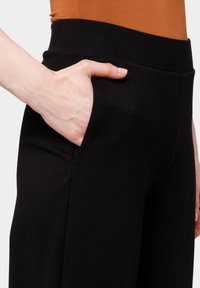 Czarne spodnie charakteryzują się wysokim stanem i kieszenią boczną, mają gładką fakturę i dopasowany krój. Pomarańczowa bluzka jest częściowo widoczna powyżej.