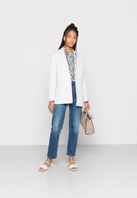 Blazer cinza claro sobre uma camisa floral branca, emparelhado com jeans azuis e sandálias de bloco bege. Ela segura uma carteira marrom clara.