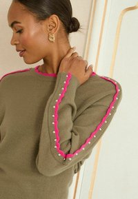 Olíva zöld pulóver, rózsaszín, hullámos díszítéssel az ujjakon, ezüst gyöngyökkel díszítve. Puha anyag és laza fazon.