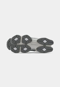 Semelle de chaussure de sport noire et grise présentant un design en caoutchouc épais avec des motifs de tread multidirectionnels et des accents blancs rectangulaires.