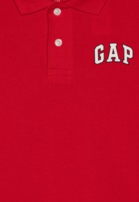 Rdeča polo majica iz teksturirane tkanine, z trakom na tri gumbe in izvezenim logotipom "GAP" v beli in črni barvi na levem prsnem delu.