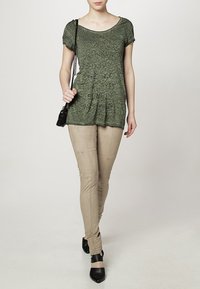 Camiseta de manga corta verde oliva con tejido texturizado y diseño gráfico en textoa, combinada con pantalones beige ajustados y zapatos de tacón negros.