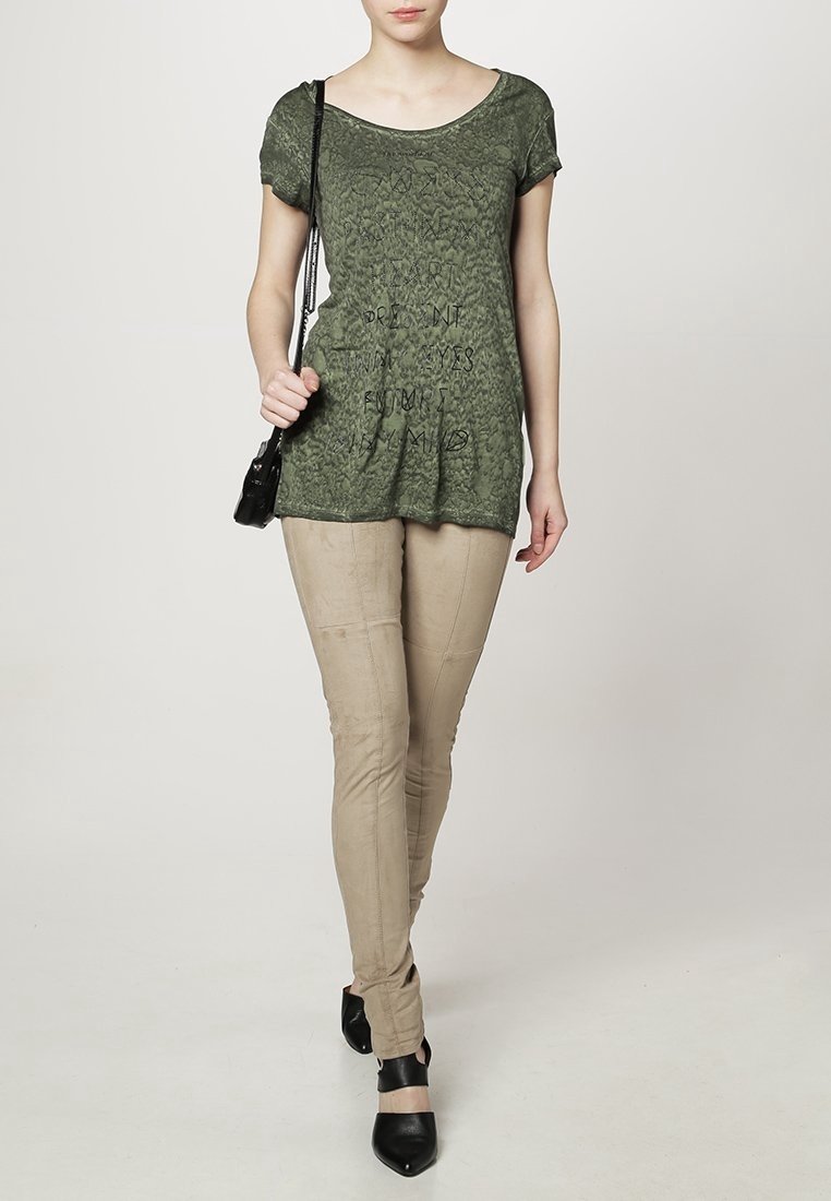 Camiseta de manga corta verde oliva con tejido texturizado y diseño gráfico en textoa, combinada con pantalones beige ajustados y zapatos de tacón negros.