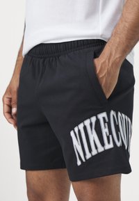 Shorts de sport noirs avec une taille élastique, arborant le logo "NIKECOURT" en broderie blanche audacieuse sur le côté.