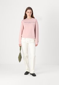 Růžová bavlněná mikina s logem "TOMMY HILFIGER EST. 1985", zkombinovaná s bílými kalhotami a zelenou taškou, postavená na jednobarevném pozadí.