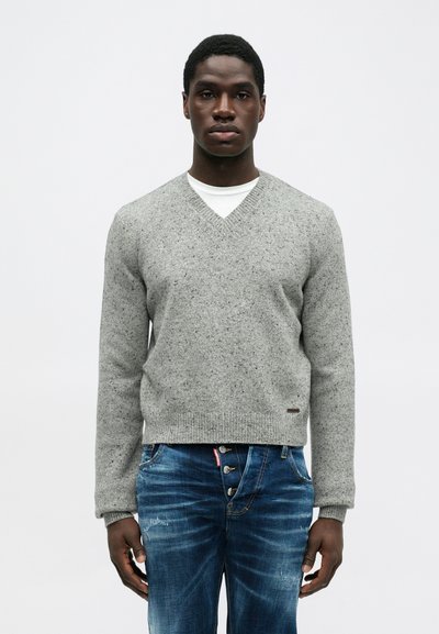 Man in een grijs gemêleerd V-halstrui over een wit shirt en blauwe versleten jeans met blootgestelde knopen, staande tegen een effen achtergrond.