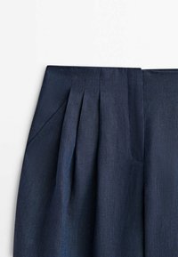Gros plan d’un pantalon taille haute bleu marine foncé sur mesure avec plis à l’avant et une poche latérale.