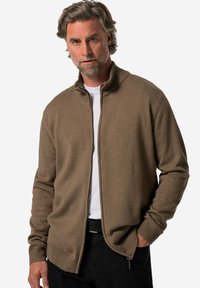 Cardigan marrón de punto con cremallera en la parte delantera, cuello acanalado y puños. Lleva una camiseta blanca debajo, cuenta con bolsillos laterales y un corte relajado.