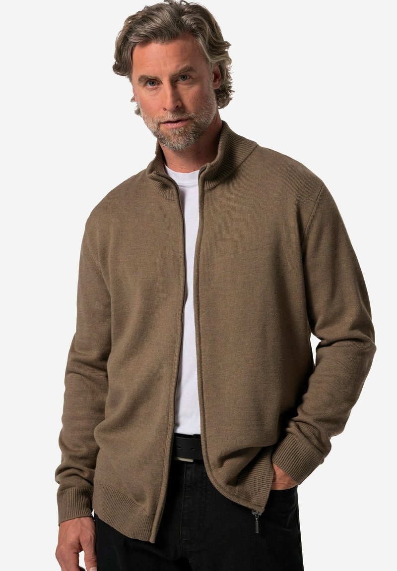 Cardigan marrón de punto con cremallera en la parte delantera, cuello acanalado y puños. Lleva una camiseta blanca debajo, cuenta con bolsillos laterales y un corte relajado.