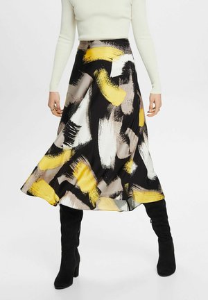 Kobieta ubrana w czarną spódnicę midi z żółtymi, białymi i beżowymi abstrakcyjnymi wzorami pociągnięć pędzla, kremową bluzkę z długim rękawem oraz czarne botki na obcasie.