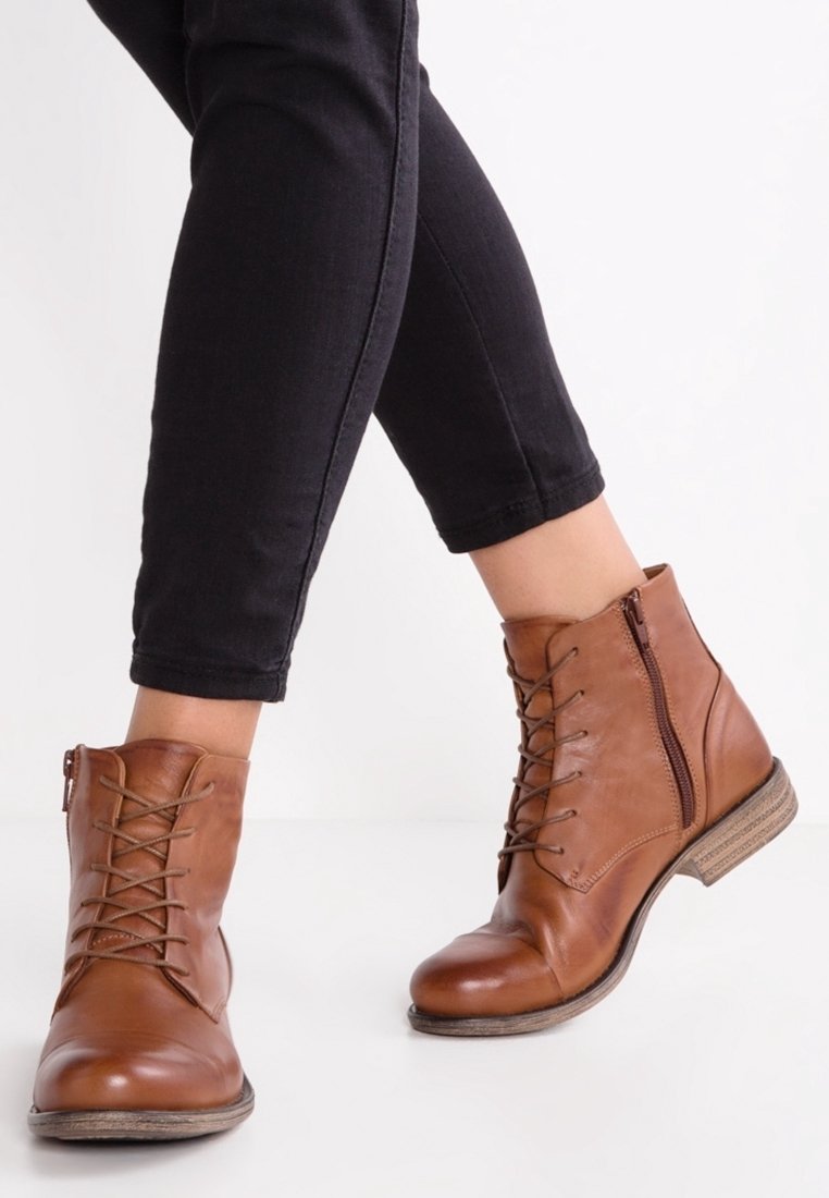 Bottes basses à lacets en cuir marron avec fermeture éclair latérale portées avec un pantalon noir ajusté, présentées sur des jambes croisées sur un fond blanc uni.