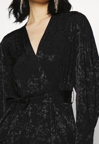 Svart blommig omlotklänning med djup V-ringning, långärmar, texturerat tyg och ett matchande bälte i midjan. Subtil lyster och elegant mönster.