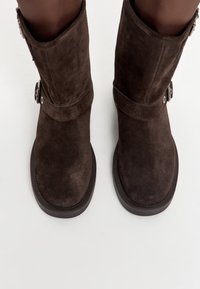 Ghete scurte din piele intoarsă maro, cu vârf rotund, talpă plată din cauciuc și accente metalice argintii pe lateral. Detaliu reglabil cu cataramă.