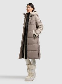 Abrigo largo acolchado en tono taupe claro, con textura acolchonada, cremallera negra y capucha forrada con piel sintética crema. Combinado con botas aislantes blancas.