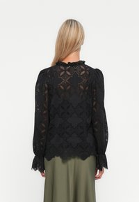 Femme aux longs cheveux blonds portant un blouse en dentelle noire avec des motifs floraux et des poignets à volants, associée à une jupe verte olive, de dos.