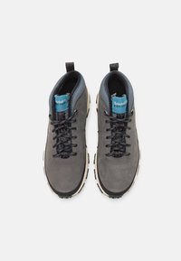 Zapatos de senderismo de cuero gris con cordones negros, un cuello azul y suelas beige. Presenta una etiqueta en la lengüeta que muestra el logotipo de la marca.