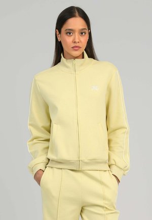 Femme portant une veste zippée jaune clair et un pantalon assorti, les mains dans les poches, debout devant un fond gris uni.