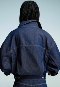 Denim jakna v temno modri barvi s sproščenim krojem, velikim ovratnikom in vidnimi šivi. Ima gumb za zapiranje v pasu.