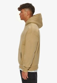 Beige hoodie with a spacious hood, kangaroo pocket, ribbed cuffs, and a smooth texture. Side view highlights relaxed fit and minimal design.