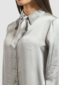 Blusa in satin grigio chiaro con colletto classico, fiocco al collo e chiusure a bottone sul davanti. Tessuto liscio e cadente.