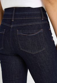 Jeans azul oscuro de tiro alto con un diseño ajustado, costuras visibles en contraste naranja y dos bolsillos traseros con detalles sutiles.