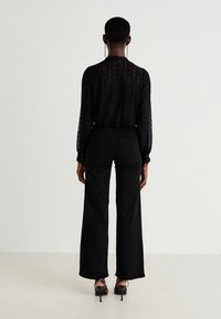 Blusa de encaje negro con mangas largas y puños con botón, combinada con pantalones de pierna ancha negros de cintura alta y tacones abiertos negros.