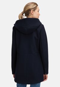 Kvinde med blondt hår i en hestehalefletning, iført en marineblå hættefrakke og blå jeans, set fra ryggen mod en simpel baggrund.
