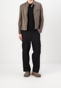 Hnědá lehká bunda se zapínáním na zip, černé tričko, černé cargo kalhoty a béžová texturovaná obuv s robustní podrážkou.