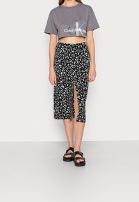Grå kortärmad t-shirt med Calvin Klein-logotyp, tillsammans med en svart midi kjol med blommönster som har en sidoöppning och svarta sandaler.