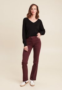 Schwarzes Langarmoberteil mit V-Ausschnitt und Knopfdetails an den Ärmeln, kombiniert mit burgunderfarbenen, engen Jeans und weißen Turnschuhen. Einfache, lässige Look.