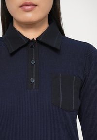 Marineblå ribbestrikket polo-skjorte med svart krage, tre knapper og en lomme med blå stripete detaljer.