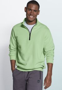 Light green pullover with a half-zip and ribbed cuffs, worn over a white shirt, paired with gray sweatpants featuring side zippers.