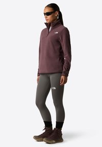 Bordeaux fleece pullover met ritskraag. Grijze leggings met logo en bruine wandelschoenen met zwarte accenten. Zonnebril gedragen.