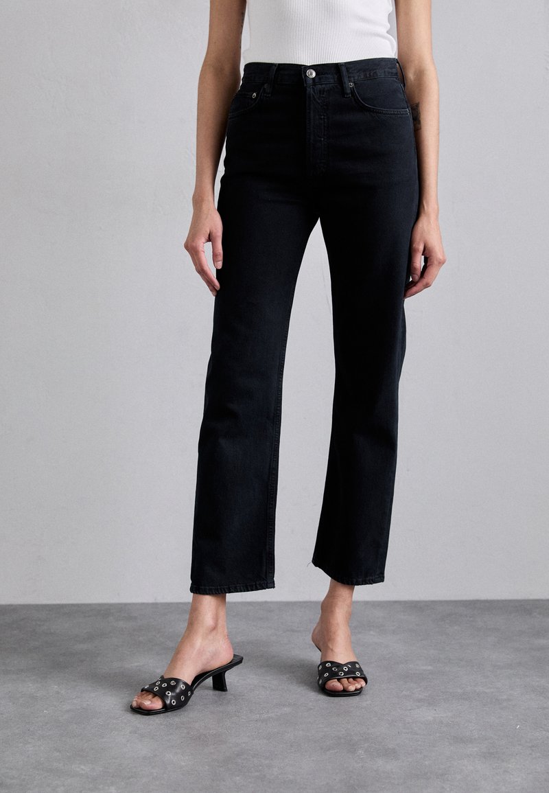 Black high-waisted jeans with a straight leg, featuring a classic five-pocket design and a button closure. Paired with black slide sandals.