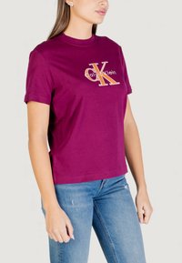 Camiseta de algodón burdeos con cuello redondo y mangas cortas, con un logo "Calvin Klein" en blanco y naranja en el frente.