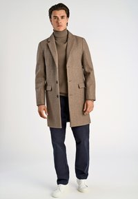 Manteau en laine marron avec revers notés, deux boutons et poches latérales, porté sur un col roulé beige, associé à un pantalon bleu marine et des baskets blanches.