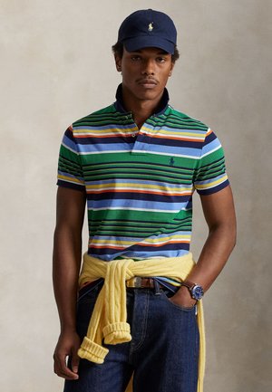 Νέος άνδρας που φοράει πολύχρωμο ριγέ πόλο πουκάμισο, κίτρινο πουλόβερ δεμένο στη μέση, σκούφο ναυτικού χρώματος, μπλε τζιν και μπλε ρολόι χειρός, στέκεται με το ένα χέρι στην τσέπη.