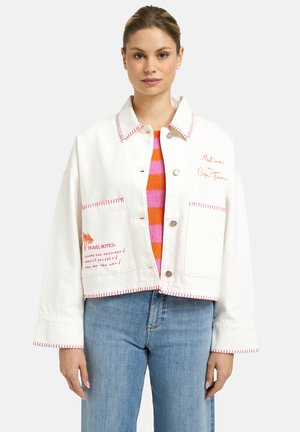 Woman wearing a white jacket with red stitching, text embroidery, and large pockets over a pink and orange striped top and blue jeans.