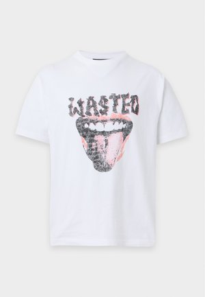 Млад мъж, носещ бяла тениска с "wasted" и графика на устни, светлосив широк панталон и черни маратонки, стои с ръце в джобовете.