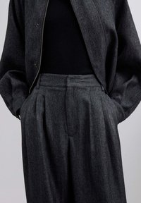 Chaqueta y pantalones de patrón espiga en gris oscuro, con un corte relajado, pliegues visibles y cierre frontal con cremallera.