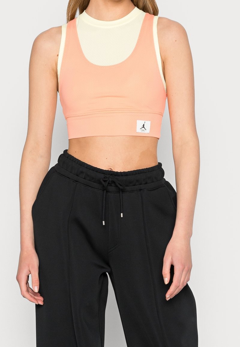Brzoskwiniowy stanik sportowy z teksturowanej tkaniny, z wysokim dekoltem i siatkowym wykończeniem. W połączeniu z czarnymi spodniami dresowymi o wysokim stanie z dodatkowymi sznurkiem.
