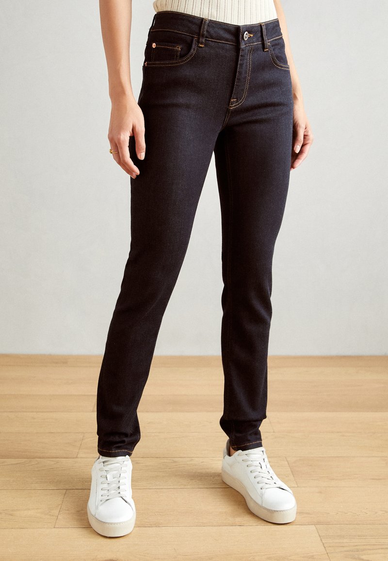 Mörka jeans i skinny-modell med kontraststickningar, fem fickor och medelhög midja, kombinerat med vita sneakers. Slät textur.