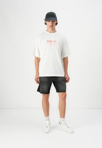 T-shirt en coton blanc avec "REPLAY" en orange, associé à un short en jean foncé. Des baskets blanches et une casquette noire complètent le look décontracté.