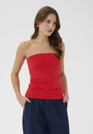 Femeie cu păr lung și maro, purtând un top roșu fără bretele și pantaloni albastru închis, stând cu mâinile în buzunare, uitându-se ușor spre dreapta.