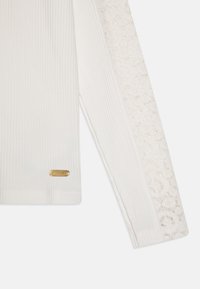 Haut à manches longues blanc avec une texture côtelée et un motif en dentelle sur une manche. Accents en métal doré près du poignet.