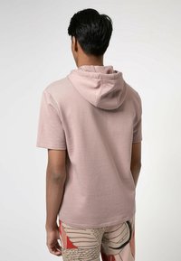 Uomo visto da dietro che indossa una felpa con cappuccio a maniche corte di un leggero mauve e pantaloni con una fantasia astratta beige, rossa e nera.