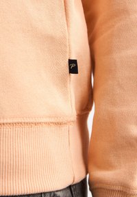 Sweat-shirt pêche avec poignets et ourlet côtelés, arborant une petite étiquette noire avec un "P" blanc sur le côté. Texture douce en coton, coupe décontractée.