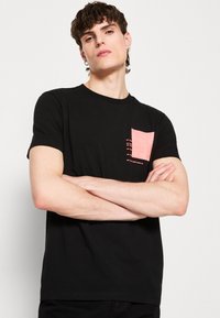 T-shirt noir en coton avec un col rond. Présente une poche rose texturée sur le côté gauche avec des accents de lignes horizontales.
