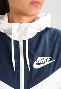 Nike vindjacka med marinblå och vit färgschema, dragkedjestängning, justerbar huva och framträdande logotyp.