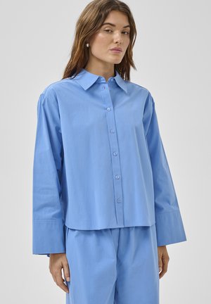 Vrouw met lang bruin haar, gekleed in een losvallend lichtblauw overhemd met knopen en bijpassende broek, tegen een effen achtergrond.